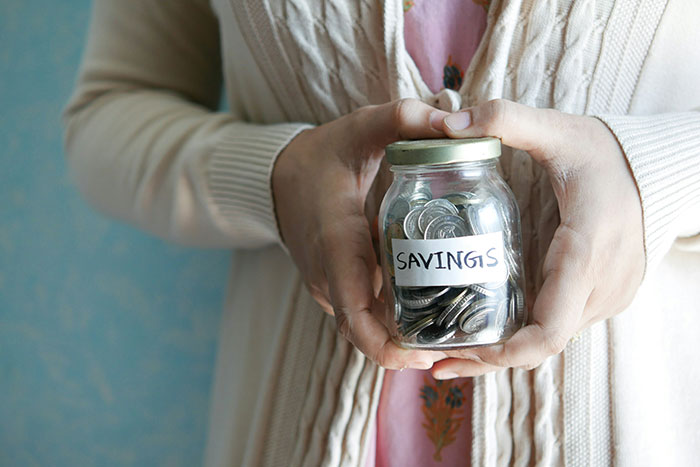 Person holding a jar labeled "savings" filled with coins, emphasizing frugality.