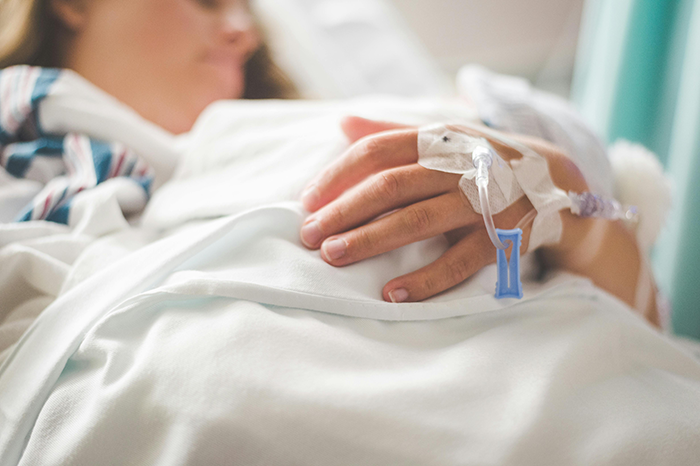 Person resting in a hospital bed with an IV, hand visible, highlighting vendor-come-wedding-deposit context.