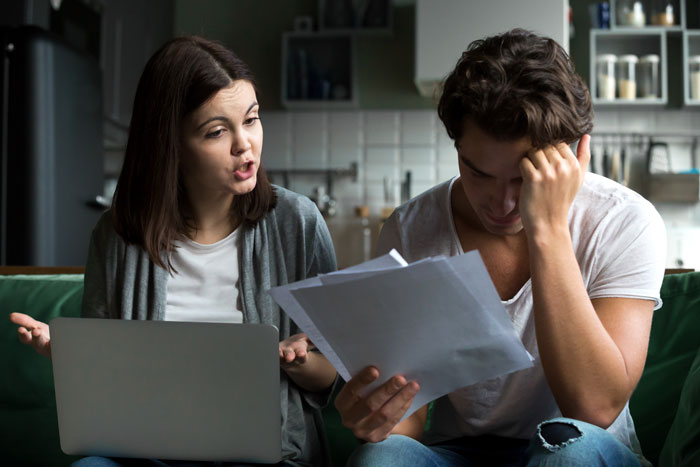 A couple having a serious discussion about finances, with a woman using a laptop and a man holding documents.