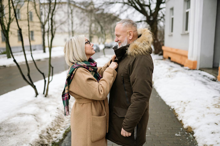 Elderly couple outdoors in winter coats, adjusting each other&rsquo;s collars, on a snowy street.