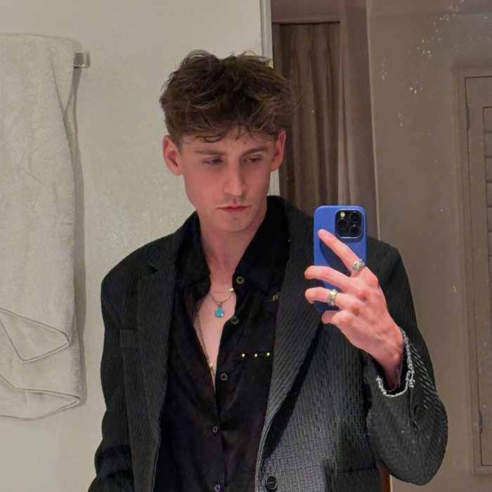 Young man in a black shirt and blazer taking a mirror selfie, smiling subtly, embodying the 'Emergency Contact' trend.