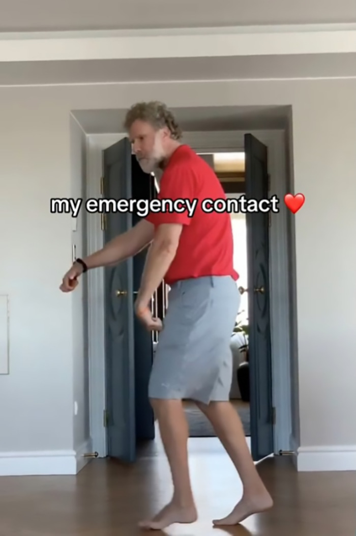 Man in a red shirt and gray shorts dancing humorously in a room, captioned "my emergency contact" with a heart emoji.