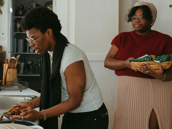 Two women in a kitchen, one with short hair and glasses, preparing food, the other holding a basket.