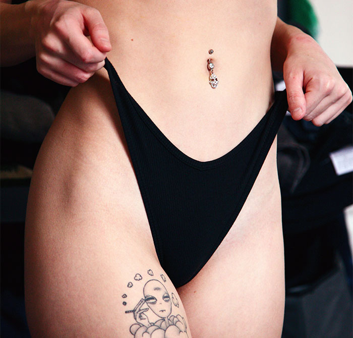 A woman with tattoo and navel piercing adjusts black underwear, related to designer genitals experience.