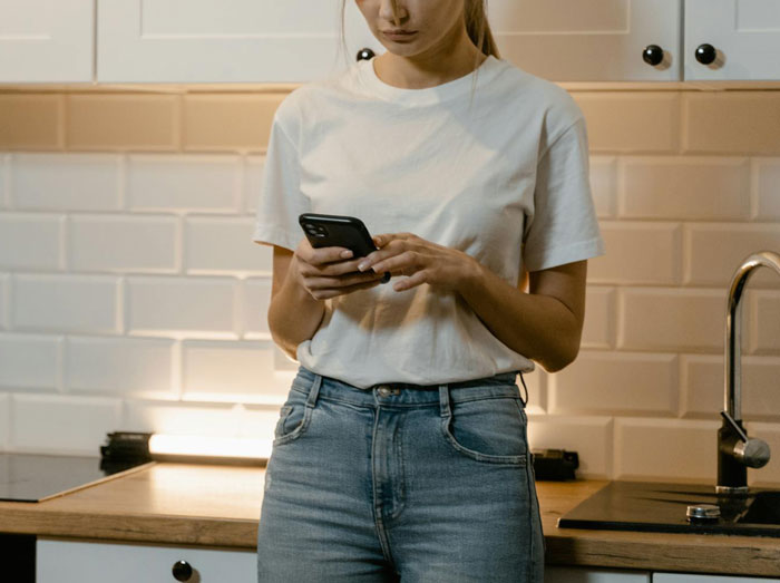 Woman in kitchen using smartphone, wearing a white shirt and jeans, related to babysitting refusal.