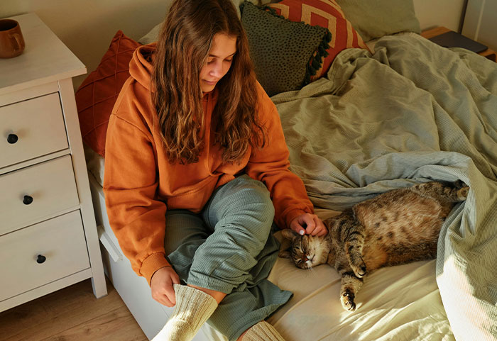 Woman in casual wear sits on bed, petting a cat, contemplating a temporary stay becoming long-term.