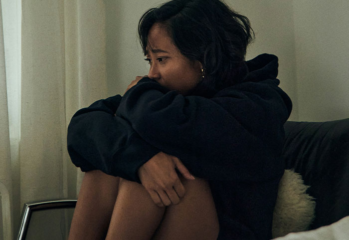 Woman sitting on a couch in a dark hoodie, knees drawn up, looking thoughtful in a dimly lit room.