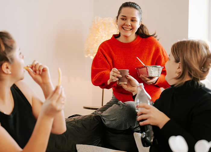 Woman in a red sweater chats with friends in a cozy room, holding a takeaway box, discussing stop gap stay.