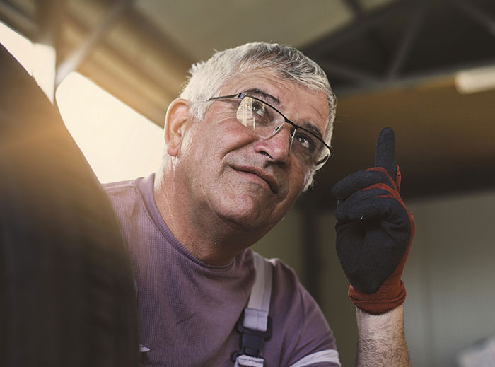 Elderly man with glasses and gloves, seemingly in thoughtful conversation, in a workshop setting.