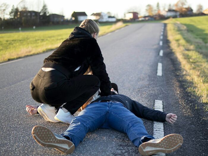 Person squatting by another lying on a rural road, sharing an unusual world perspective.