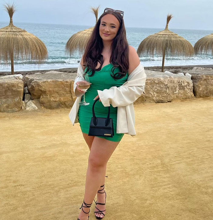 Influencer in a green dress holding a glass, standing by the beach with straw umbrellas in the background.