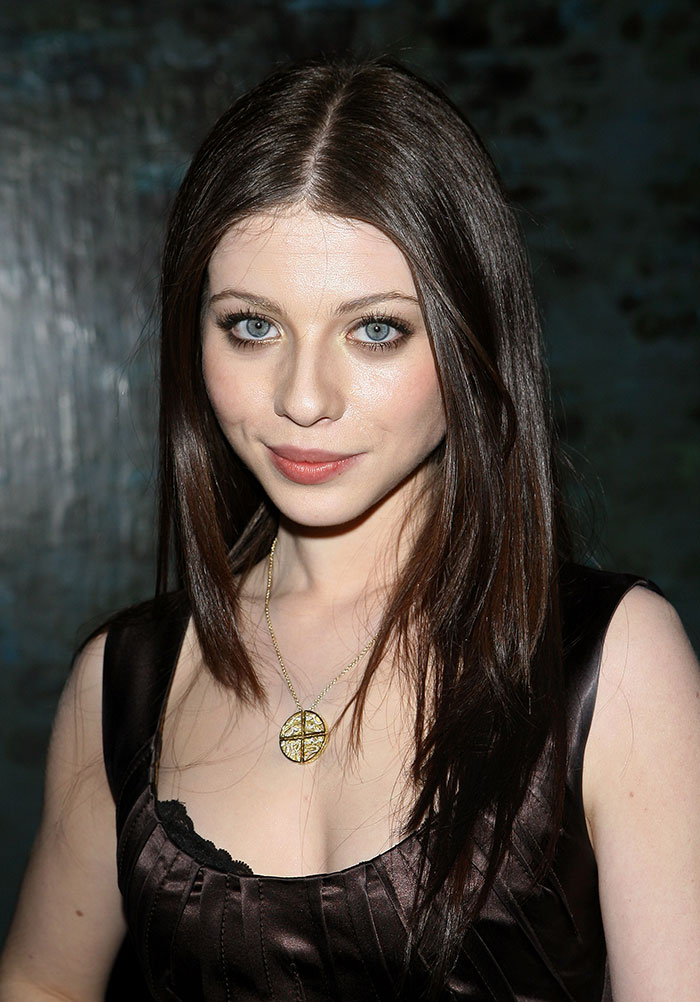 Young woman with long brown hair, wearing a pendant necklace and dark dress, posing for a photo.