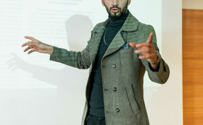 Man in a gray coat presenting in front of a projector screen, gesturing while discussing random world facts.