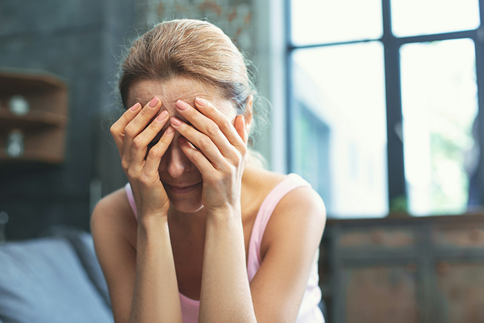 Woman in distress, sitting with hands on face, light pink top, in a dimly lit room highlighting marital issues.