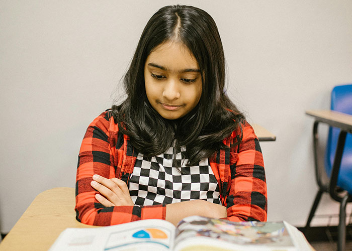 Girl sitting at a desk in a classroom, looking at a book, symbolizing a teacher pronouncing names incorrectly.