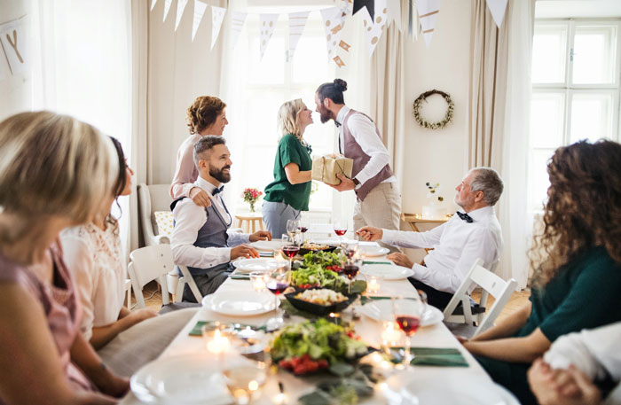 Man at rehearsal dinner table, embracing woman, surrounded by guests, with festive decor.