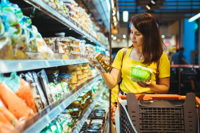 Woman shopping for cheap alternatives in grocery aisle, examining product labels.