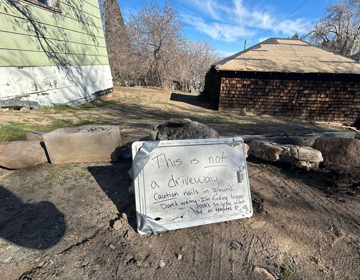 Sign reads "This is not a driveway. Caution nails in ground," warning neighbors against driving on the yard.