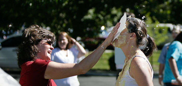 Woman playfully smashes a pie in another woman&rsquo;s face outdoors, surrounded by laughing friends.