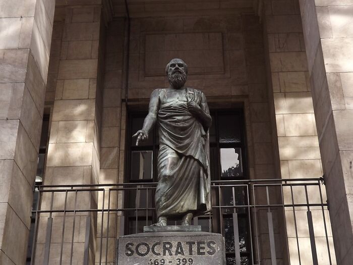 Statue of Socrates in a contemplative pose, standing on a pedestal with engraved dates at a historic building entrance.