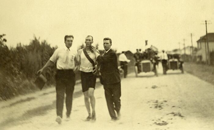 Three men on a dusty road during an early 20th-century race, with vintage cars in the background.