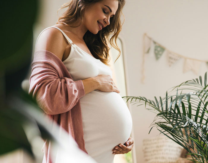 Pregnant woman smiling and holding her belly, wearing a white dress and pink cardigan, next to indoor plants.