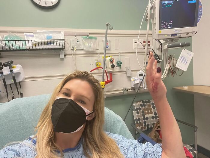 Woman in hospital bed wearing a mask, raising her left hand; medical equipment in the background.