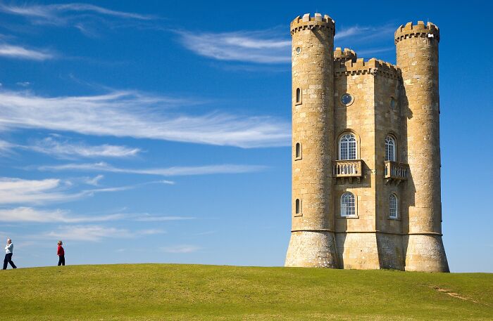 Historic stone tower with two people walking nearby under a clear blue sky.