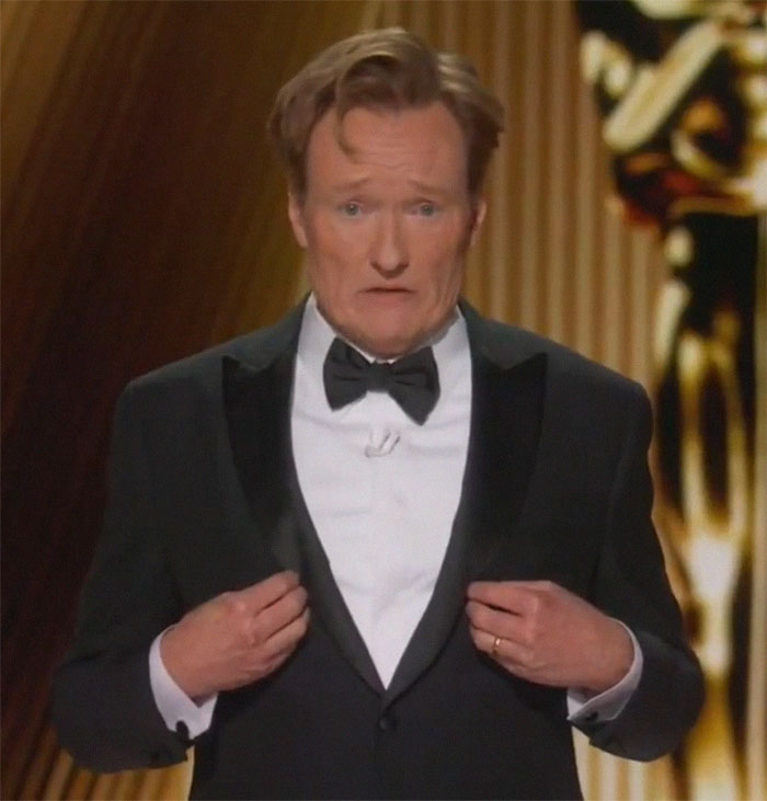 Comedian at Oscars delivering controversial jokes in a tuxedo on stage.