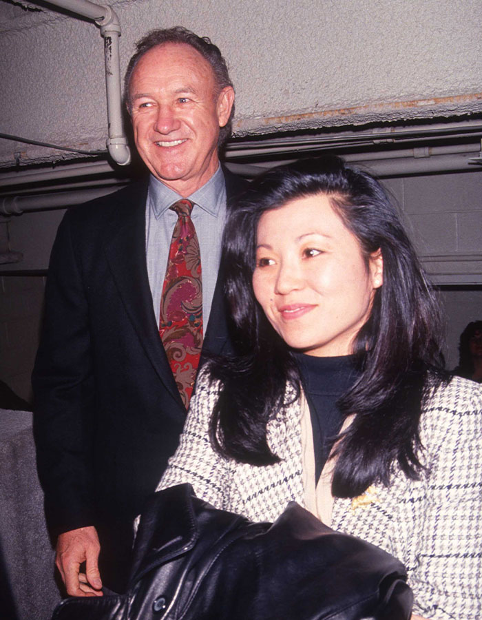 Elderly man in a suit smiling, standing next to a woman in a checkered jacket; focus on Gene Hackman topic.