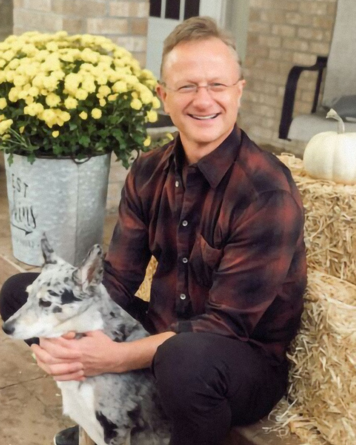 Man sitting with a dog, wearing a plaid shirt, smiling. Bright yellow flowers and autumn decor in the background.