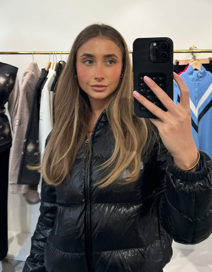 Woman in black puffer jacket taking a mirror selfie in a clothing store.