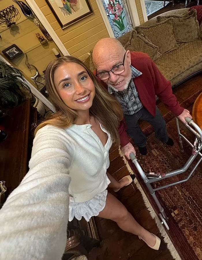 Adult star taking a selfie with an elderly man using a walker in a cozy room.