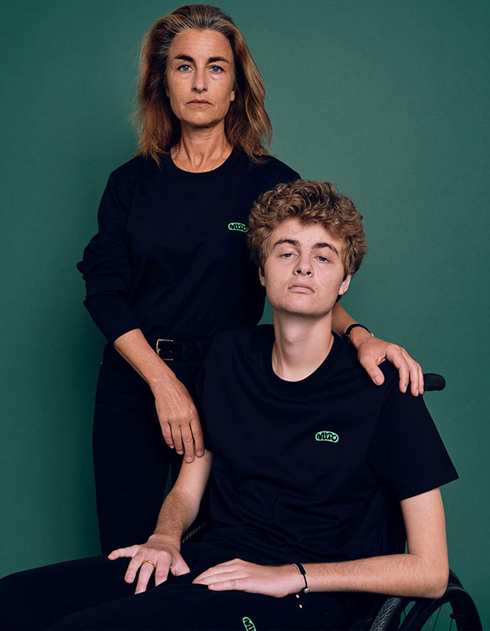 Woman standing beside a young man in a wheelchair against a green background, both wearing black shirts.