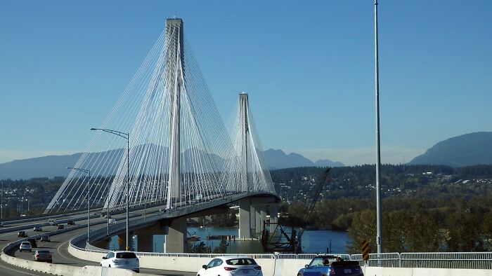 Cable-stayed bridge with cars driving, against a backdrop of mountains and a river, offering a cool architectural view.