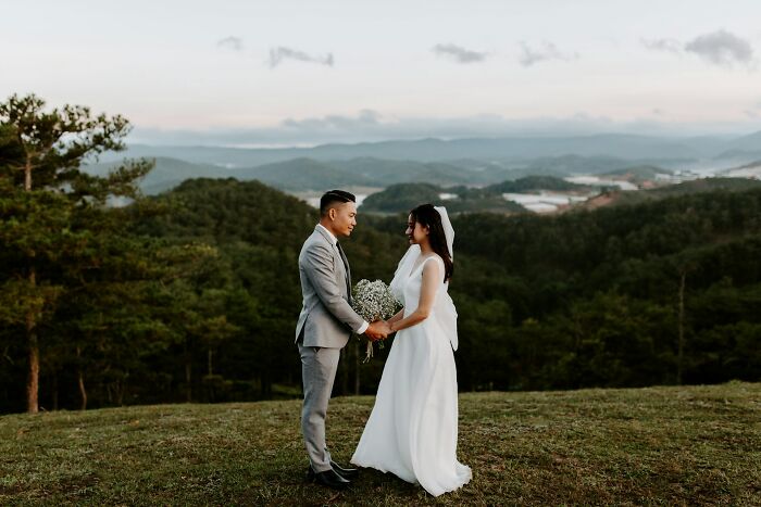 A couple in wedding attire stands outdoors, amidst scenic hills, facing each other.