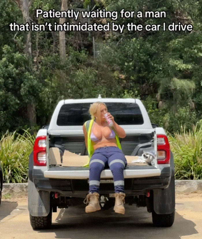 Woman in a bikini top and work gear, sitting on truck, advocates for working topless like male co-workers on hot days.