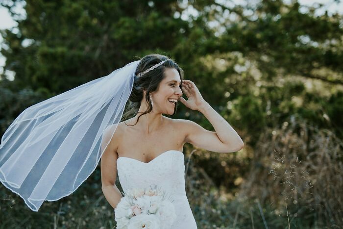 Bride in a white dress and veil smiling outdoors, holding a bouquet.