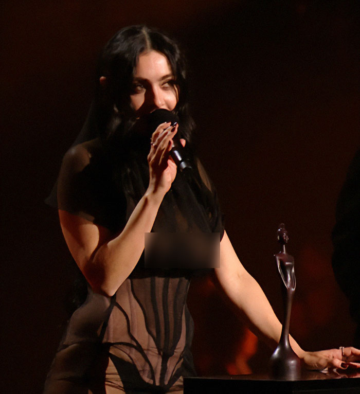 Charli XCX wearing a sheer dress on stage with a microphone in hand.