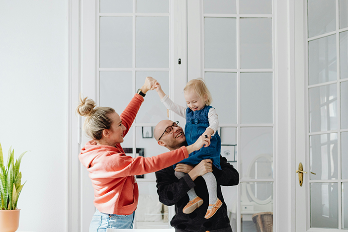 Family moment in a bright room, with parents lifting their child playfully near a large window.