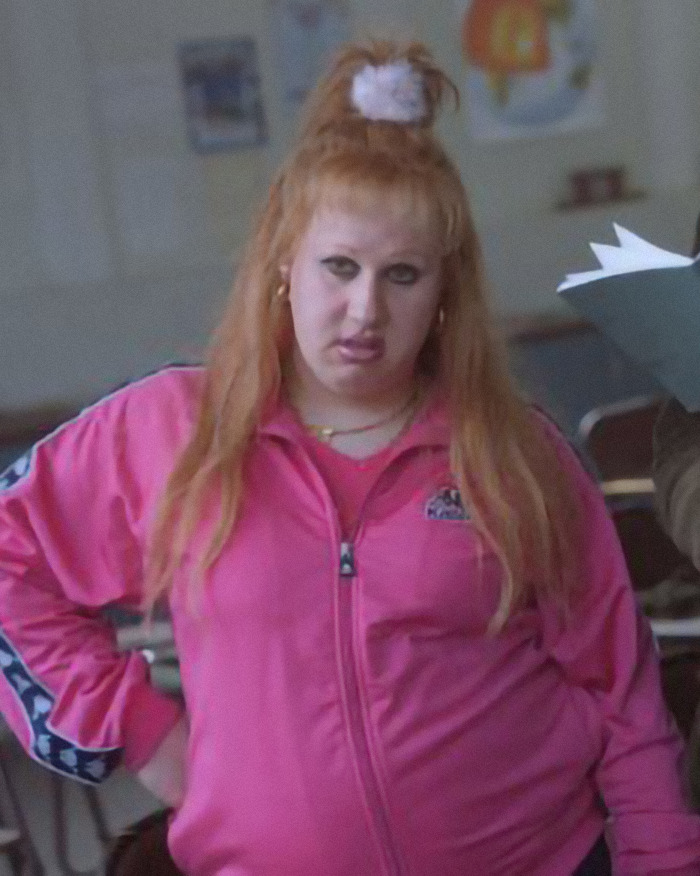 A person in a pink track jacket with long hair and scrunchie, representing the comedian involved in the "Mommy Makeover" jab.