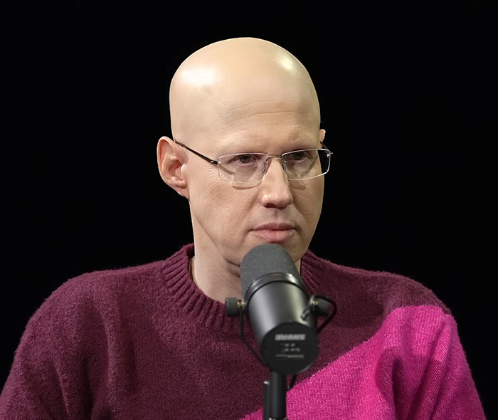 Bald comedian wearing glasses and a maroon sweater, speaking into a microphone.