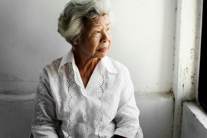 Elderly woman in white shirt gazing thoughtfully out a window, seated indoors.