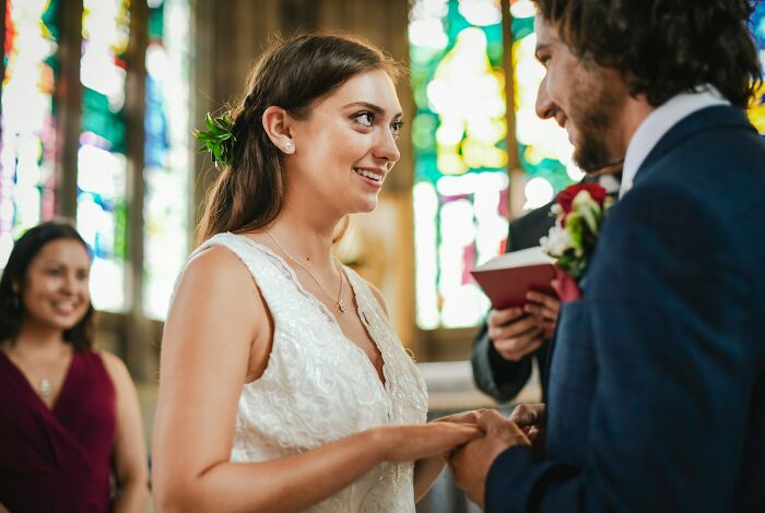 Bride and groom exchanging vows, holding hands, amid wedding guests and stained glass backdrop.