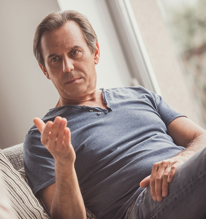 Man in a blue shirt seated on a couch, gesturing with his hand, discussing family care responsibilities.