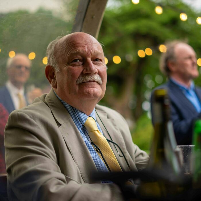 Elderly man at a wedding, seated in a beige suit with a yellow tie, surrounded by guests, under string lights.