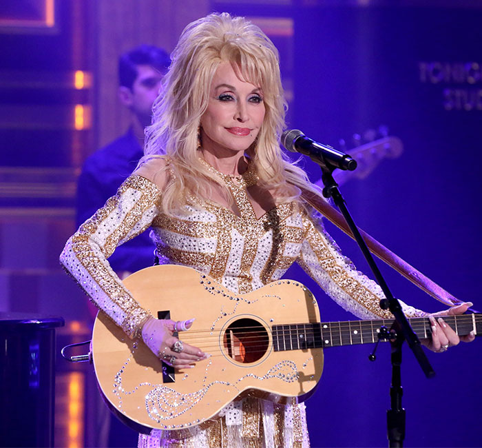 Dolly Parton performing on stage, holding a guitar, in a sparkling outfit.