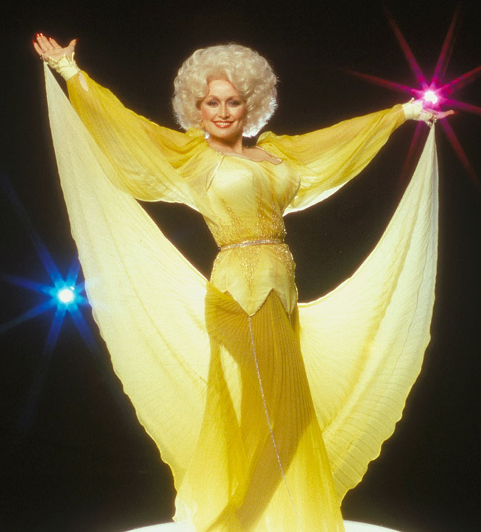 Dolly Parton in a sparkling yellow dress, arms outstretched, with colorful stage lights behind her.