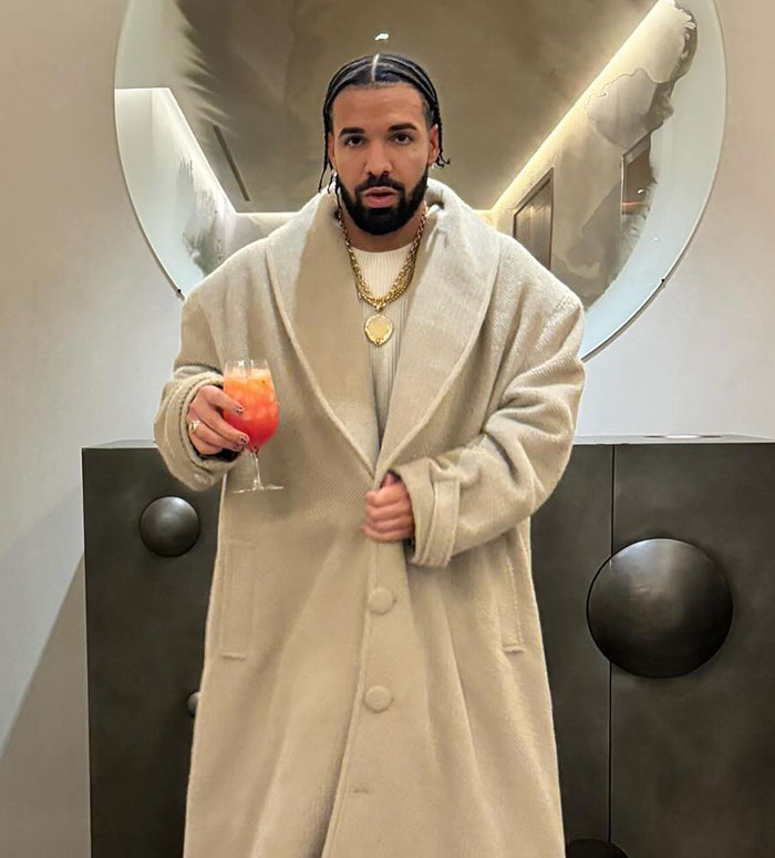 Man in a beige coat holding a drink indoors, standing with a calm expression.