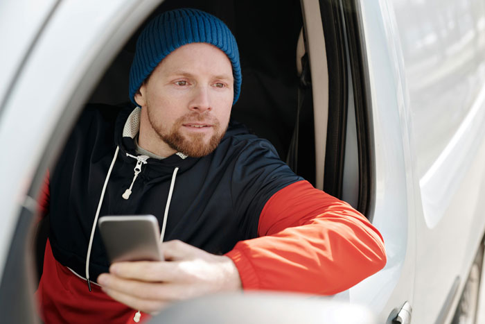 Driver in a van wearing a blue beanie, holding a smartphone, possibly planning petty revenge over tolls paid.